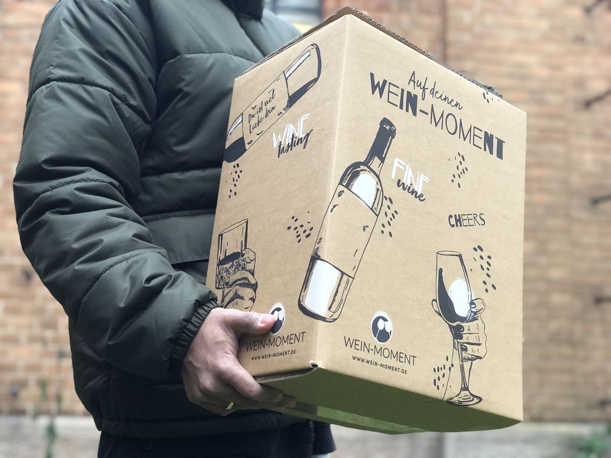 Person trägt einen bedruckten WEIN-MOMENT Versandkarton mit der Aufschrift „Auf deinen Wein-Moment“